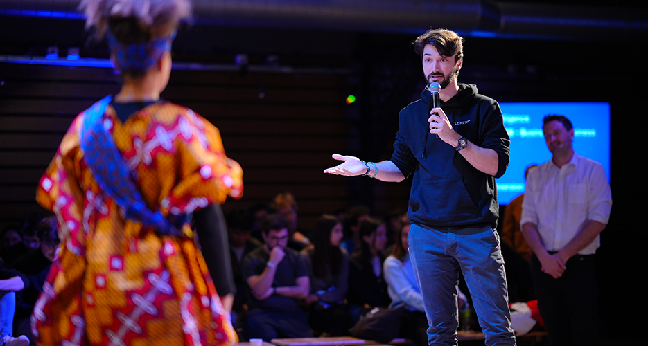 Man speaking into a microphone on stage during an indoor event, engaging with a person in colorful attire, with an audience seated in the background.