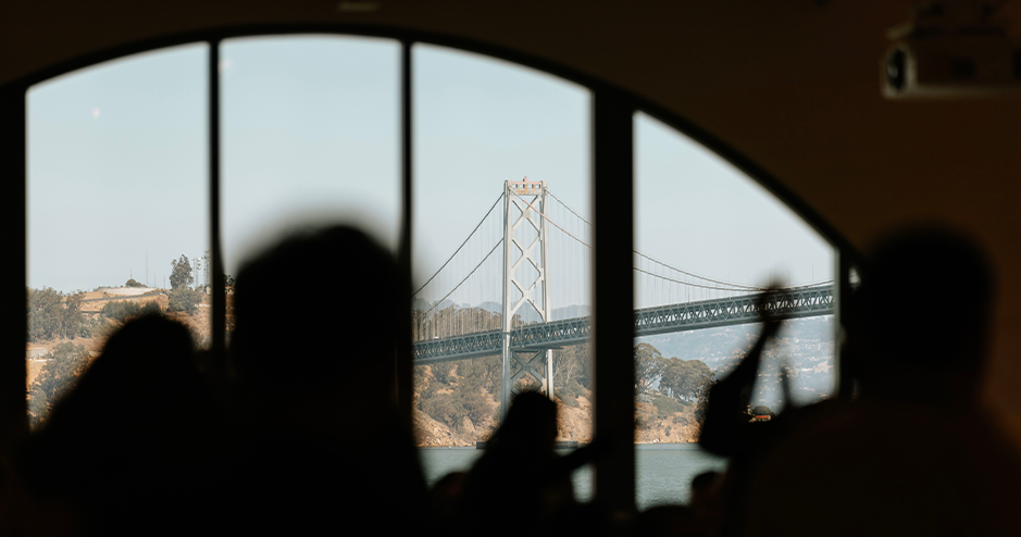 View of a suspension bridge seen through large arched windows, with the silhouette of people in the foreground. The bridge stretches across a body of water, with hills and trees visible in the background.