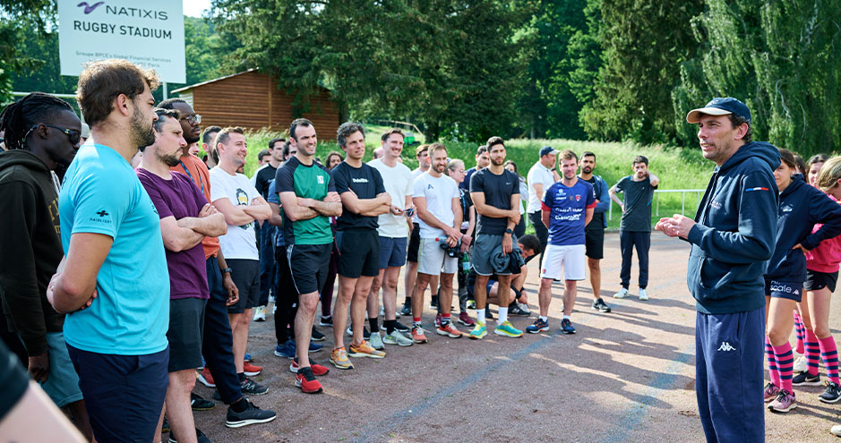 Groupe de personnes rassemblées dans un stade de rugby, écoutant quelqu'un qui leur parle devant eux