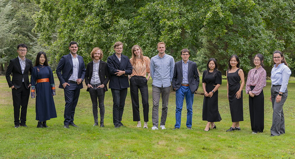 The image shows the same group of eleven individuals standing in a line outdoors on a grassy field. They are in a park-like setting, with large green trees in the background. The group is dressed in formal to semi-formal attire, with a mix of suits, dresses, and casual clothing. The individuals are smiling and appear to be posing for a professional or team photo