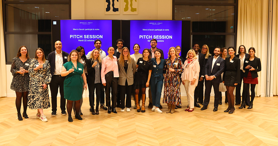 group of professionals poses together in a well-lit event space with wood flooring and modern lighting fixtures. They stand in front of two large screens displaying the text 'PITCH SESSION, Mardi 22 octobre' and 'Merci d’avoir participé à notre Pitch Session.