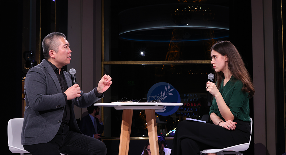 Yi Zeng in conversation with another speaker during a panel at the Paris Peace Forum, with the Eiffel Tower illuminated in the background