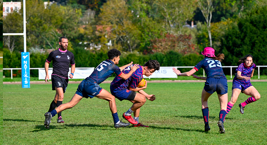 A rugby player wearing a purple jerseyHEC-Natixis Mixed Rugby Nations Cup 2024: the HEC Paris team takes on Imperial College (Student University Tournament) is tackled by an opponent in a blue jersey during an outdoor rugby match. A female player wearing a pink hat and another player are approaching the action. A referee is visible in the background. A Natixis advertising banner is placed next to the goalposts.