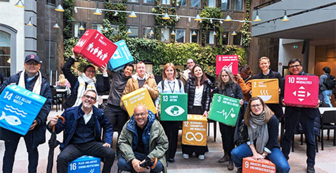 A diverse group of people posing in an indoor space with green walls, holding colorful blocks representing the United Nations Sustainable Development Goals (SDGs)