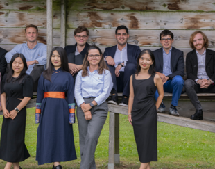 The image shows a group of eleven people posing outdoors in front of a wooden structure. Five individuals, mostly men, are sitting or leaning on the top part of the structure, while six others, mostly women, are standing in front of it on the grass