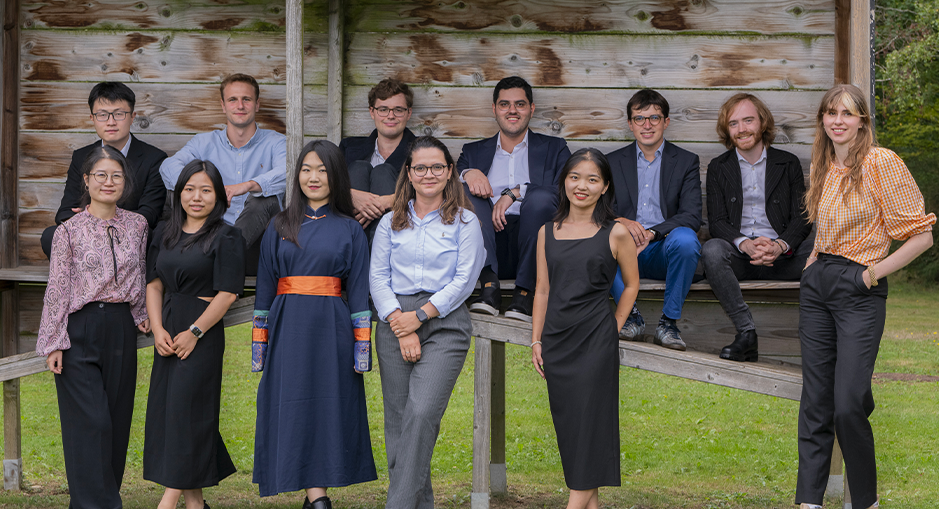 The image shows a group of eleven people posing outdoors in front of a wooden structure. Five individuals, mostly men, are sitting or leaning on the top part of the structure, while six others, mostly women, are standing in front of it on the grass