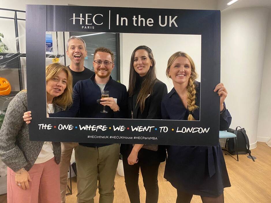 Five smiling people posing with a photo frame that reads HEC Paris In the UK - The One Where We Went to London' at an event in London