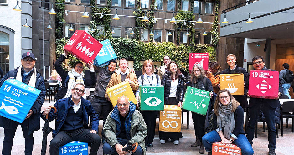 A diverse group of people posing in an indoor space with green walls, holding colorful blocks representing the United Nations Sustainable Development Goals (SDGs)